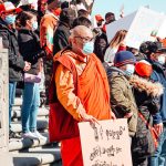 A diverse group of people, including a monk, participate in a protest holding banners and flags on a sunny day.