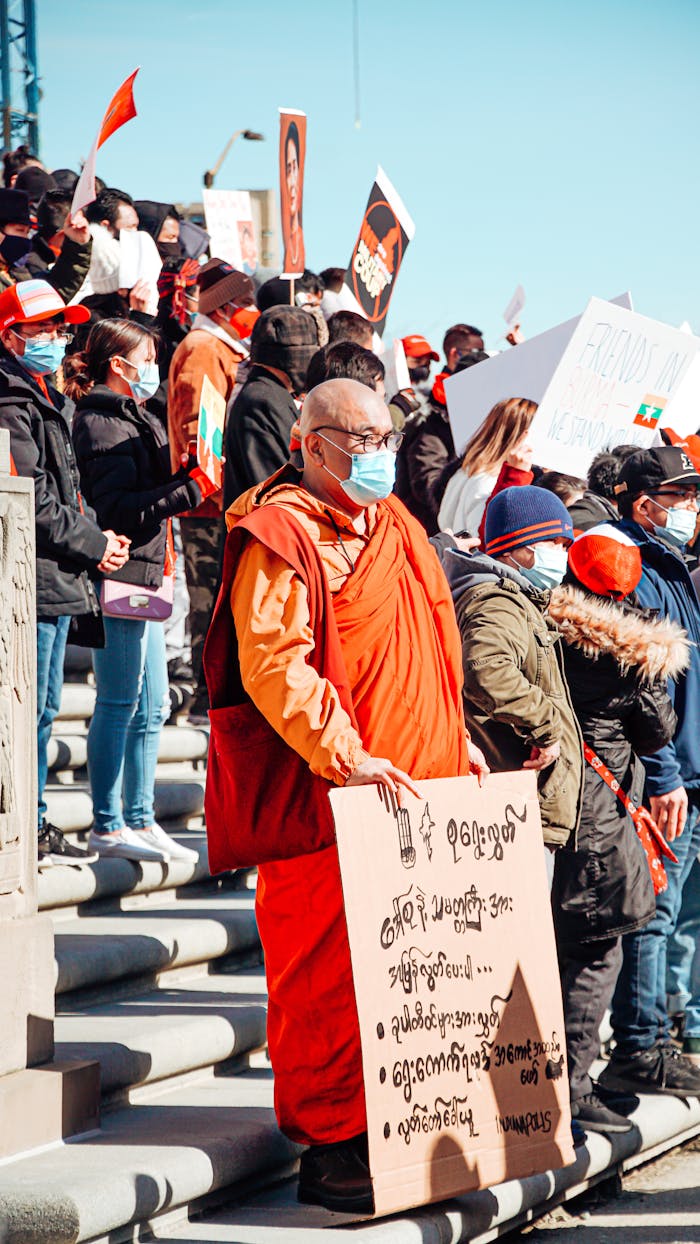 A diverse group of people, including a monk, participate in a protest holding banners and flags on a sunny day.